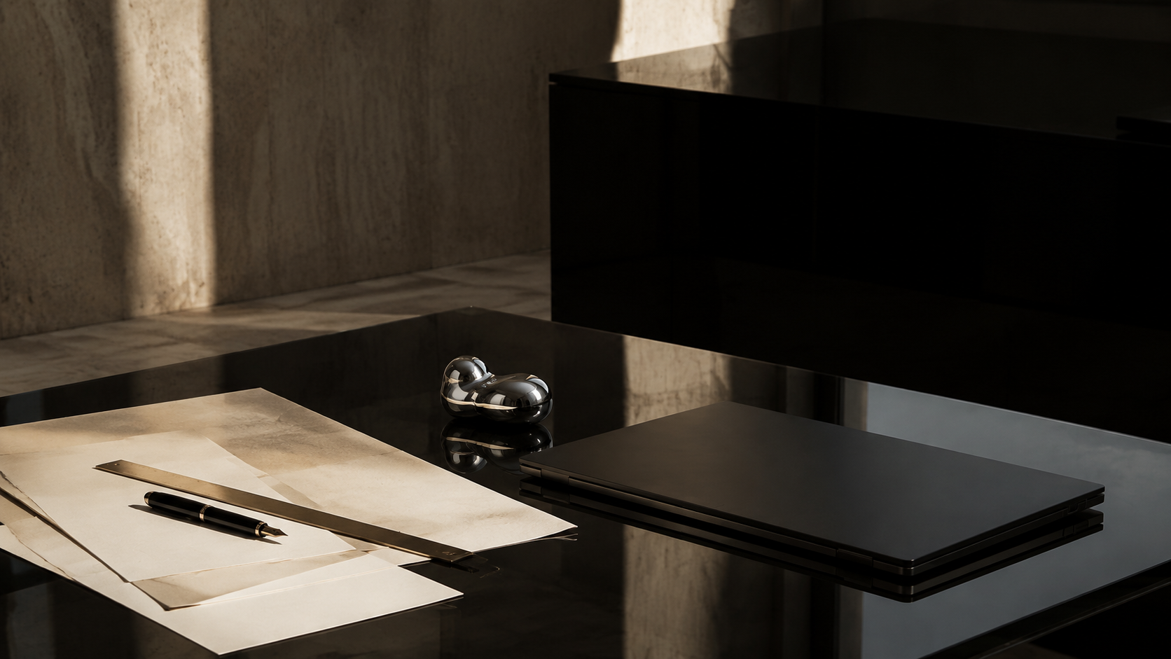 A black glass studio desk with ivory papers, a muted gold ruler, a black fountain pen and a closed black laptop in warm limestone architecture.